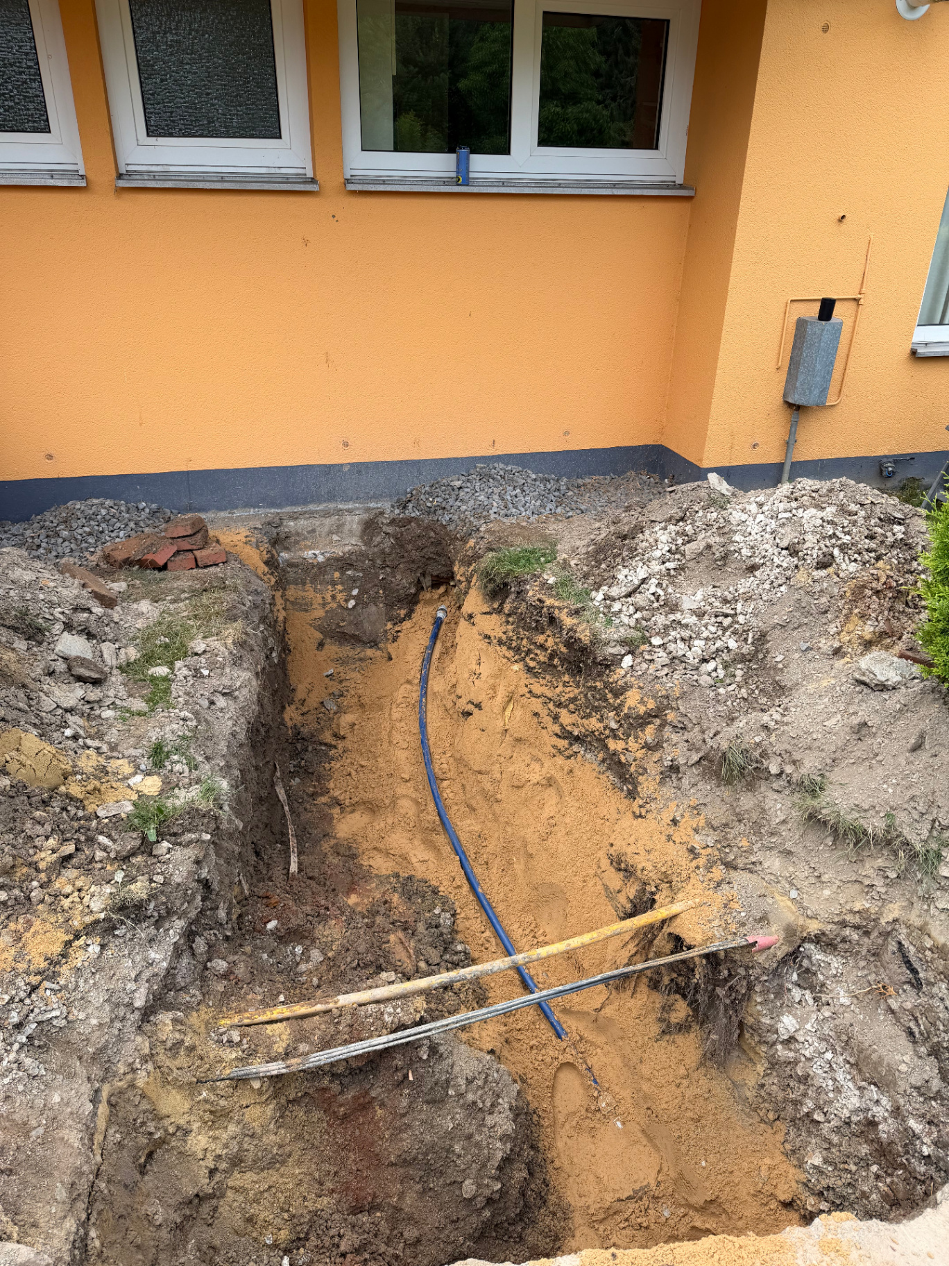 Offene Baugrube vor einem Haus mit sichtbaren Leitungen und Kabeln im Tiefbau