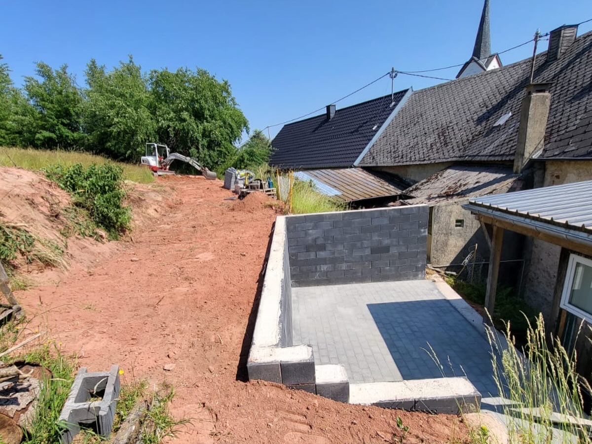 Neu erstellte Stützmauer und Pflasterfläche in Hanglage in Arenrath mit Kirchturm im Hintergrund